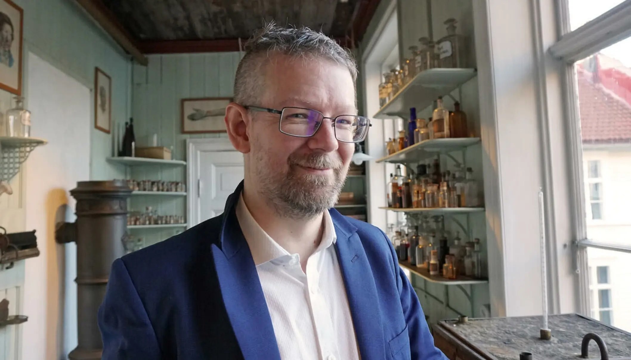 En mann med briller og skjegg, iført blå blazer og hvit skjorte, står i et gammeldags rom med flaskehyller og store vinduer.
