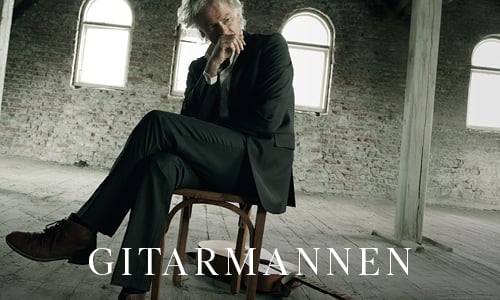 En mann i dress sitter på en trestol i et tomt, rustikt rom med buede vinduer og holder en ettertenksom positur. Ordet "GITARMANNEN" står nederst.