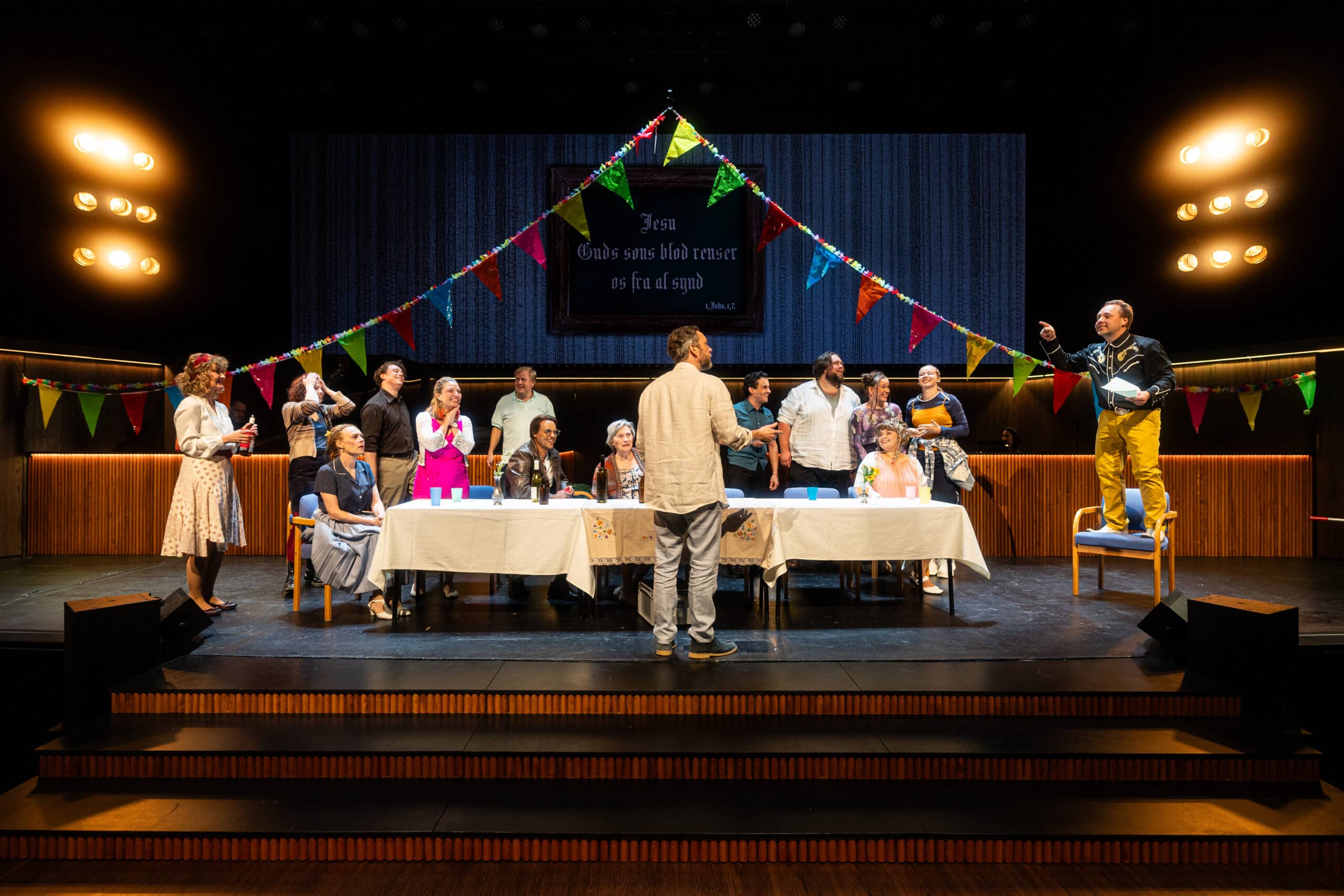 En gruppe mennesker er samlet rundt dekorerte bord på scenen under fargerike vimpler, og én person står og taler til de andre i en teatralsk forestillingssetting.