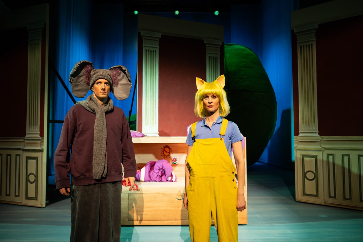 To skuespillere i dyrekostymer står på scenen; den ene har elefantører og snabel, den andre har gule katteører og kjeledress. Bak dem står en fargerik kulisse med søyler og en stor grønn figur.