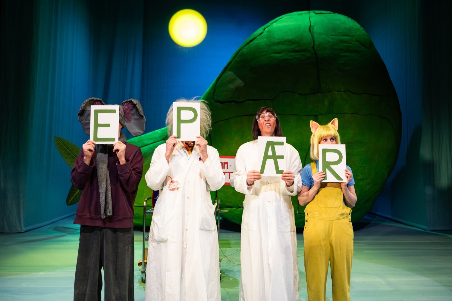 Fire utkledde personer står på scenen og holder opp skilt med bokstavene "E", "P", "Æ" og "R" foran en stor grønn rekvisitt og et skarpt lys over.