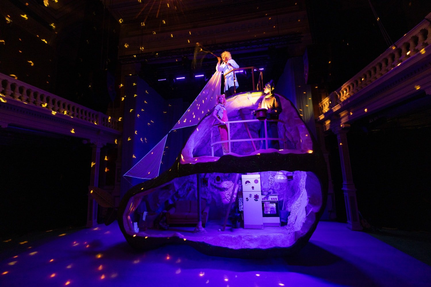 En scenografi består av en stor, tverrskåret hodestruktur med to personer inni og én person over, alt under lilla belysning med projisert gult lys.