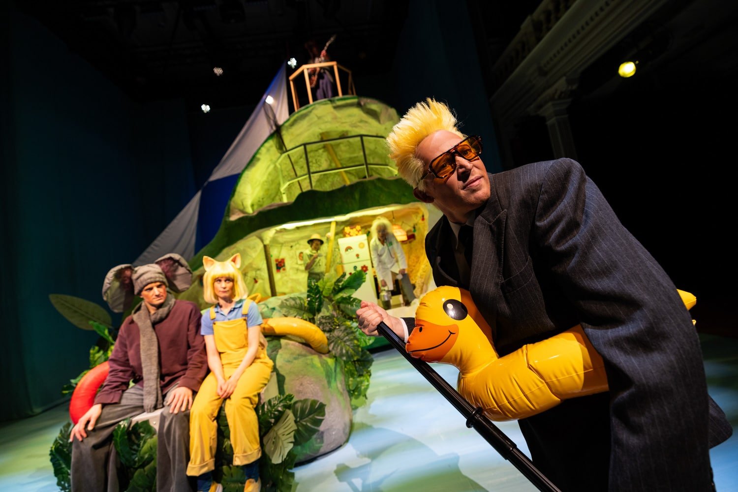 En mann i dress med spikert blondt hår, solbriller og en oppblåsbar and står foran to personer i fargerike kostymer, med en grønn, finurlig kulisse i bakgrunnen.