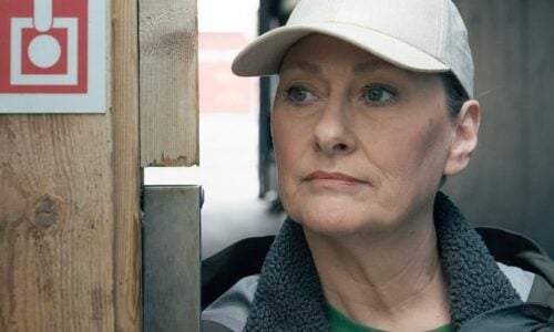 En kvinne med en lys lue og jakke står ved en vegg med et skilt som viser en nødutgang, og ser til siden. Teksten på norsk viser filmtittelen «Sønnen» og opplysninger om rollebesetningen.
