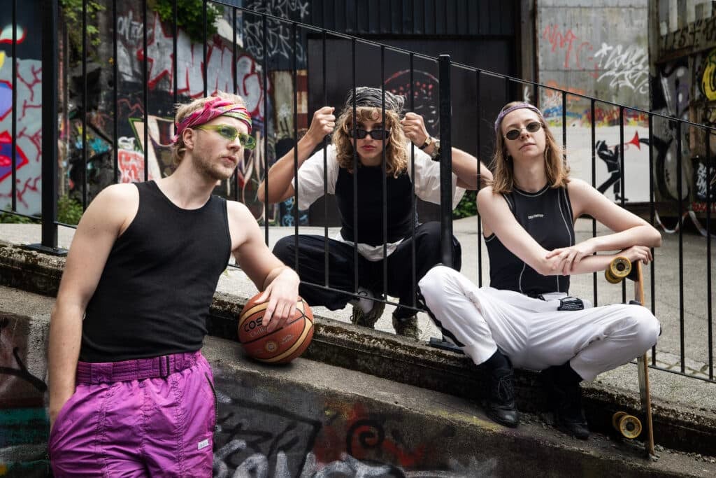 Tre unge voksne i sporty klær poserer i et urbant område med graffiti; en holder en basketball, en annen sitter på huk bak et rekkverk, og den tredje har et skateboard.