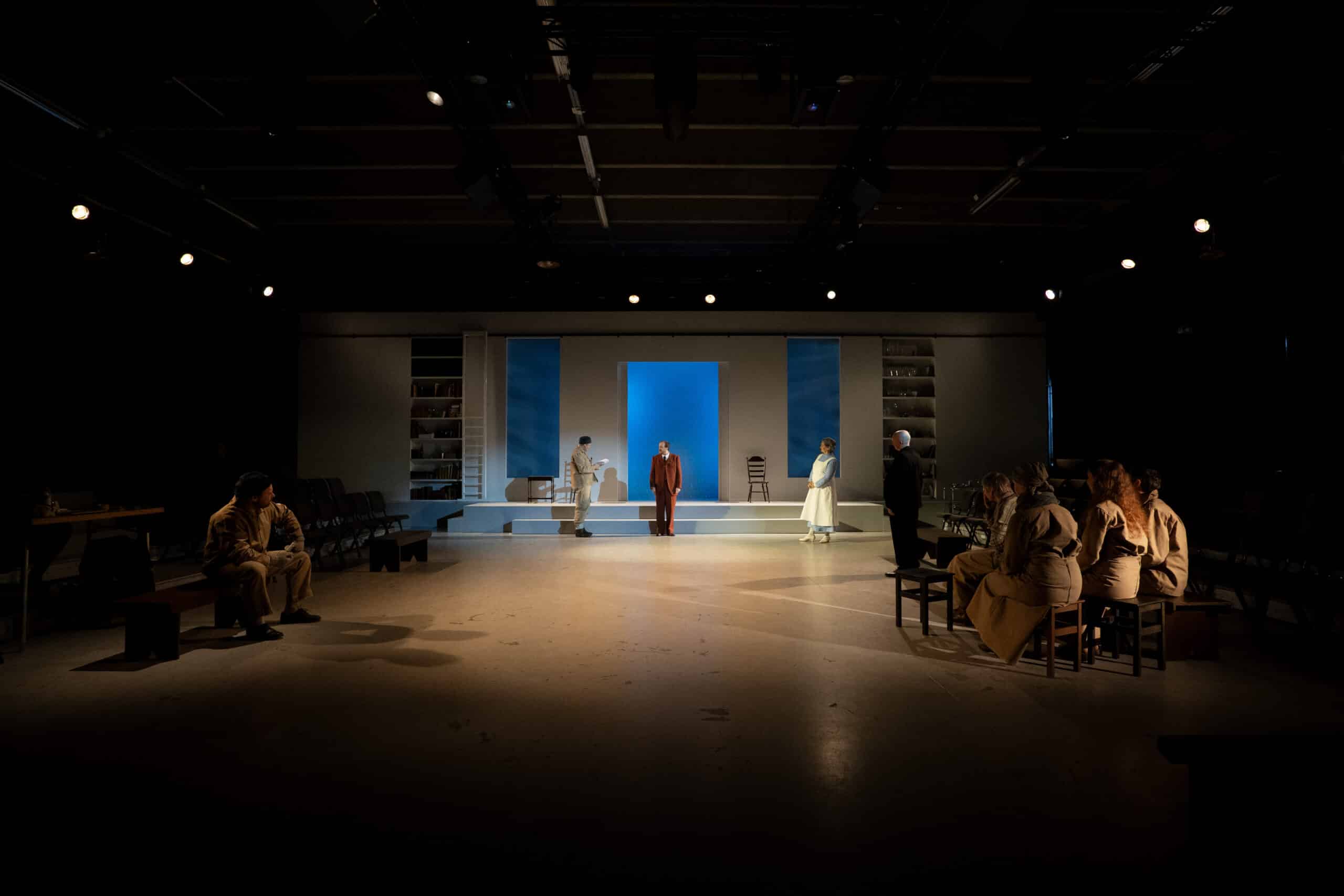 En sceneforestilling viser skuespillere fordelt over en minimalistisk scenografi med blå belysning, mens sittende publikummere ser på fra begge sider.