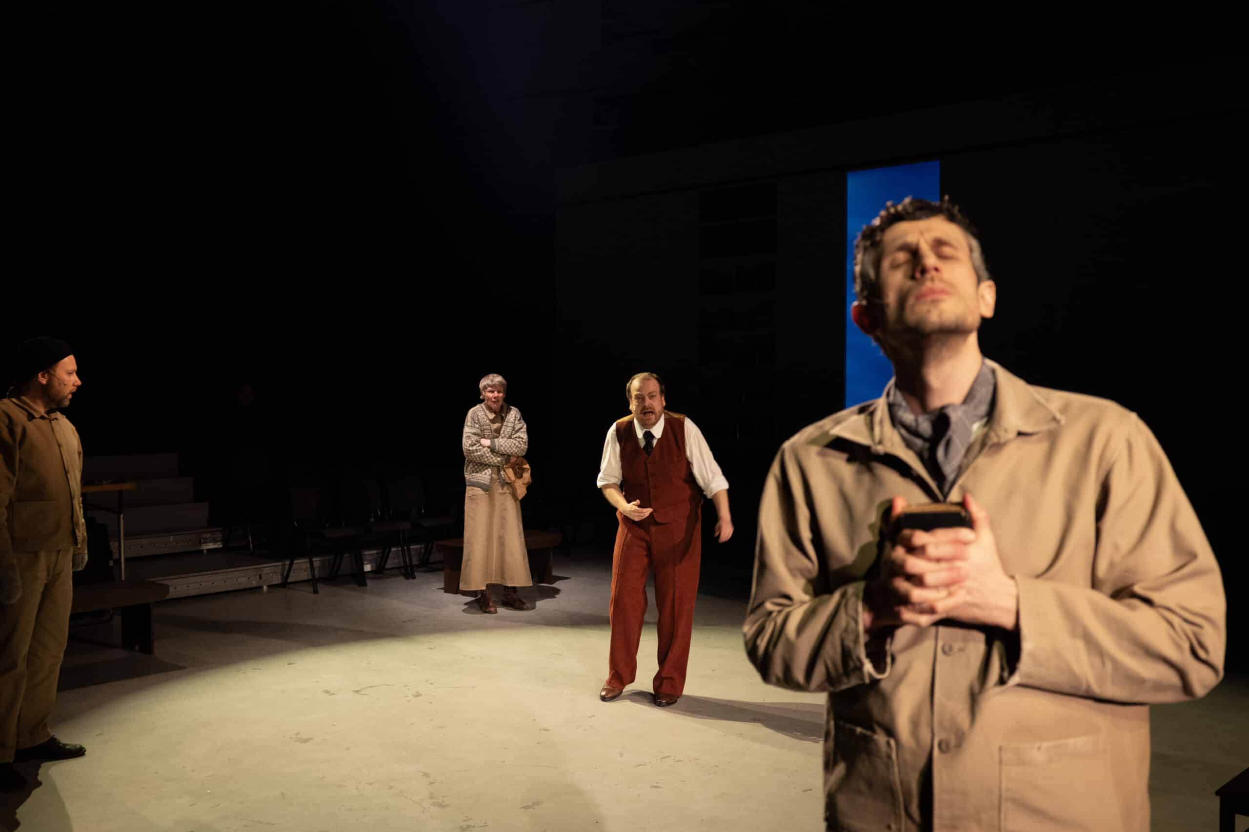 Fire skuespillere opptrer på en svakt opplyst scene; en mann står i forgrunnen og holder en gjenstand med lukkede øyne, mens tre andre engasjerer seg i bakgrunnen.