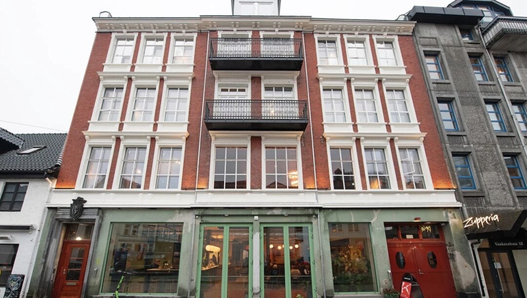 En fireetasjes bygning i rød murstein med store vinduer, grønne dobbeltdører i første etasje og balkonger i andre og tredje etasje. En grønn scooter står parkert foran bygningen.
