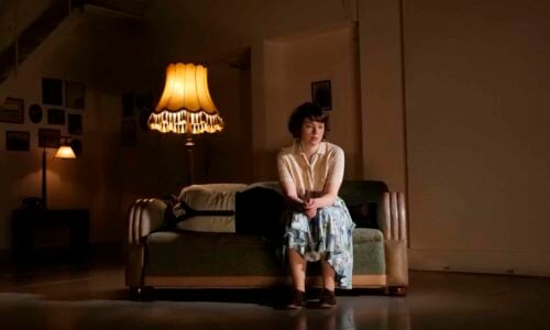 En kvinne sitter alene på en sofa i et svakt opplyst rom med vintage lamper og dekor.