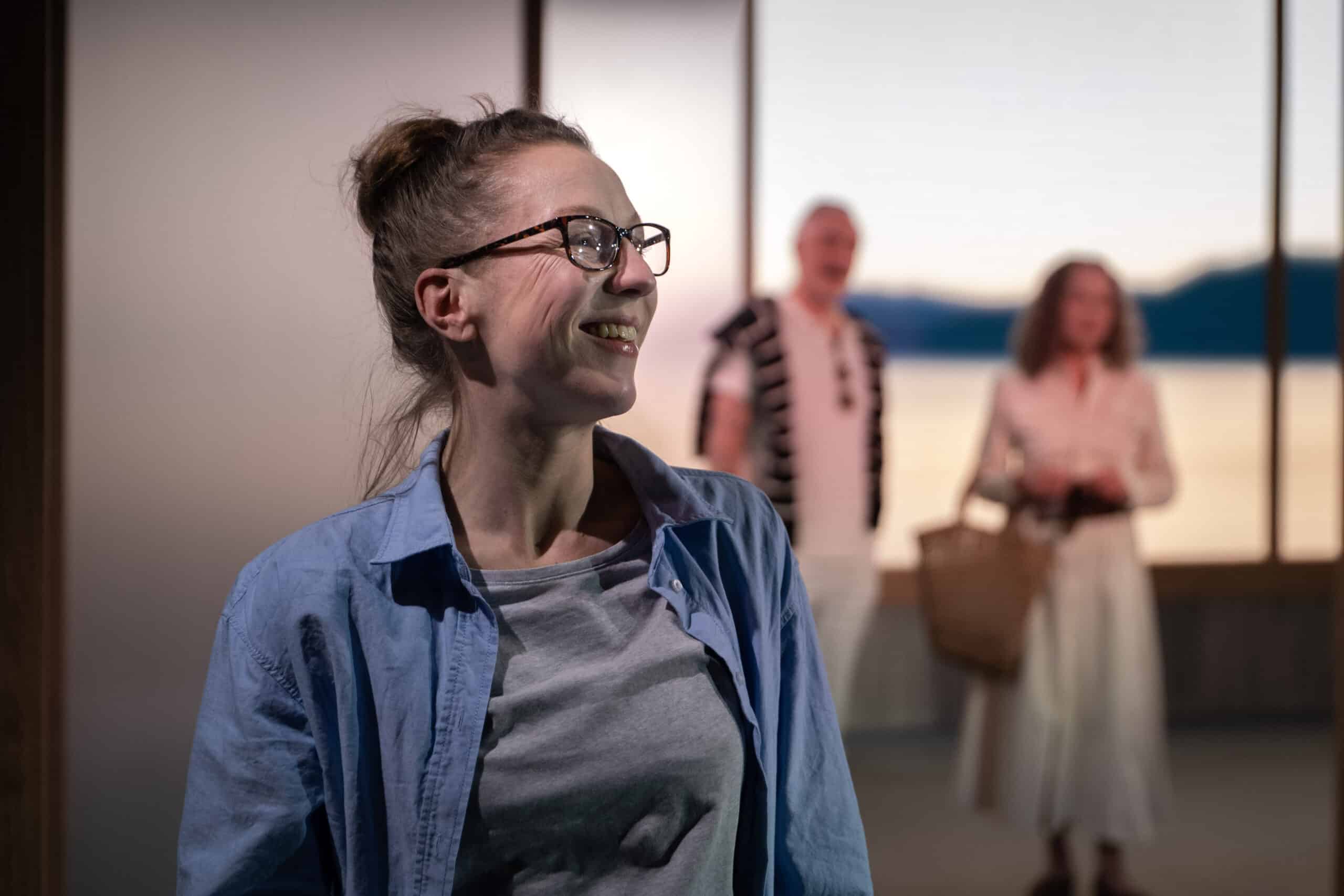 En kvinne i briller smiler i forgrunnen. To personer står i den uskarpe bakgrunnen, den ene holder en vevd pose.