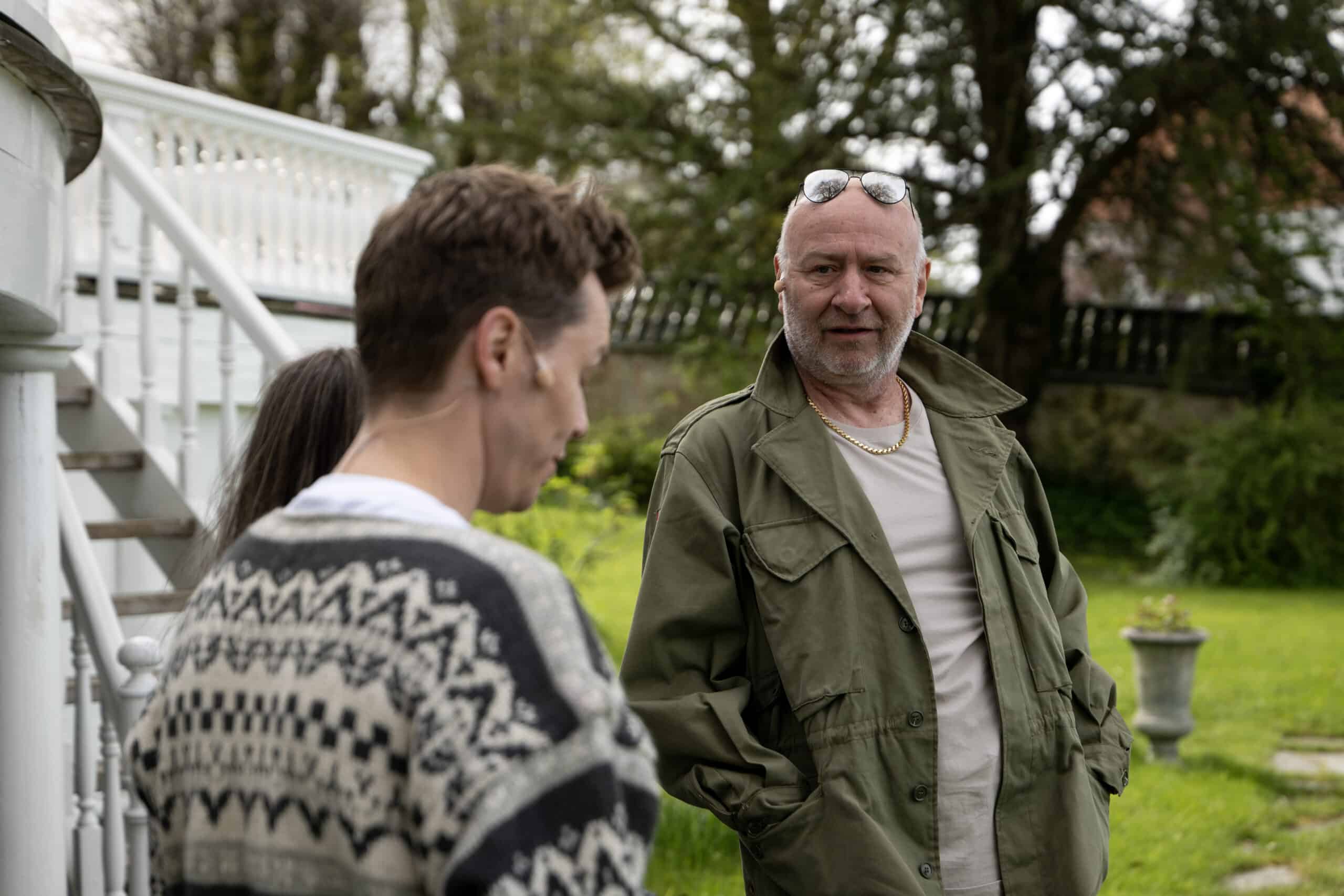 To menn står utenfor ved en trapp og snakker; den ene har på seg en mønstret genser, den andre har på seg en grønn jakke og solbriller på hodet. Trær og gress er i bakgrunnen.