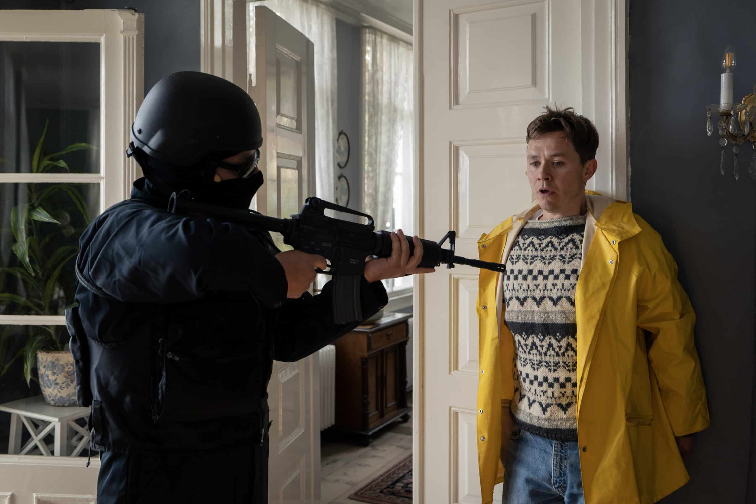 En politibetjent i taktisk utstyr retter en rifle mot en overrasket mann i gul regnfrakk som står mot en dør inne i et hus.