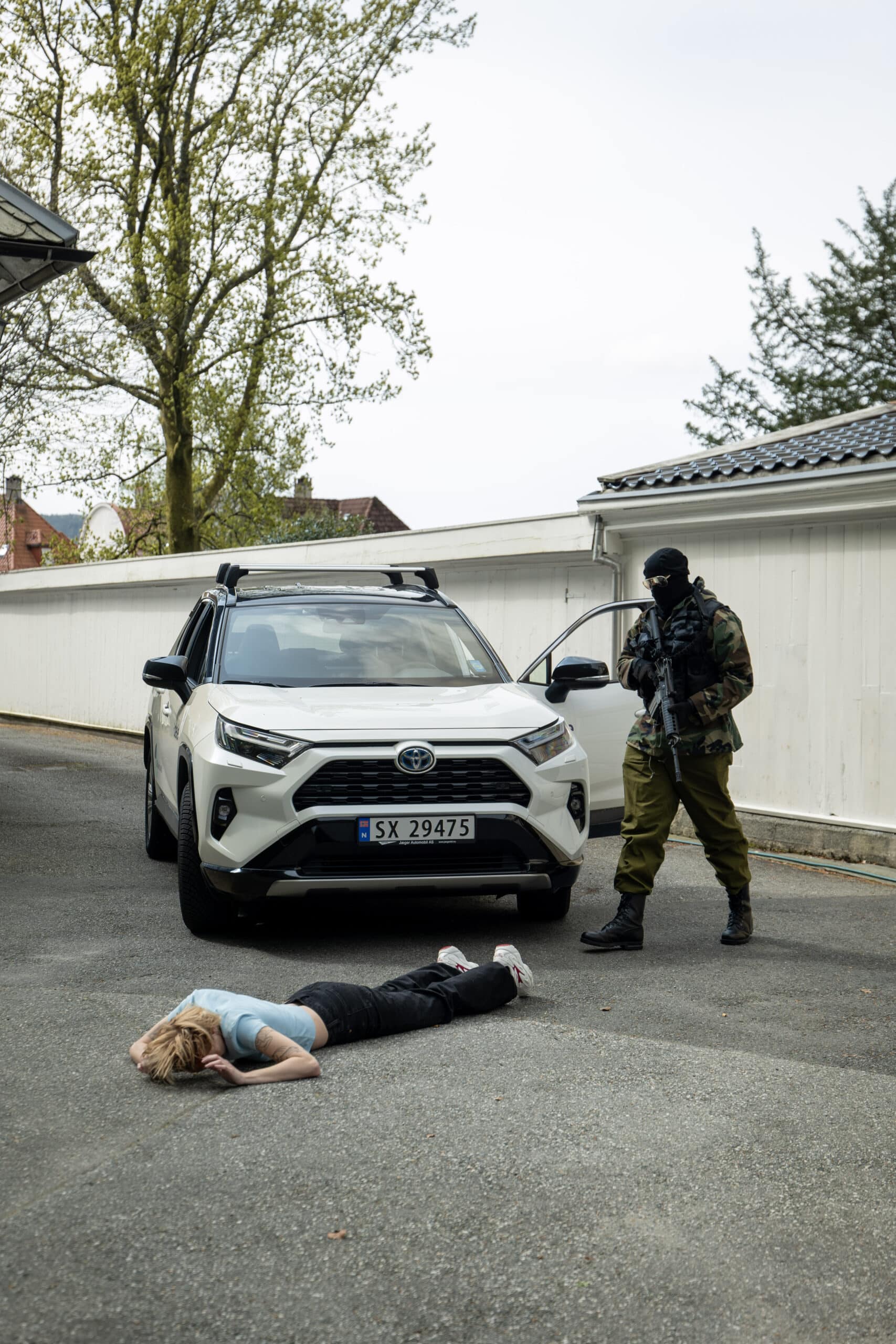 En person ligger med ansiktet ned på bakken foran en hvit SUV, mens en bevæpnet, maskert person i militærantrekk står i nærheten.