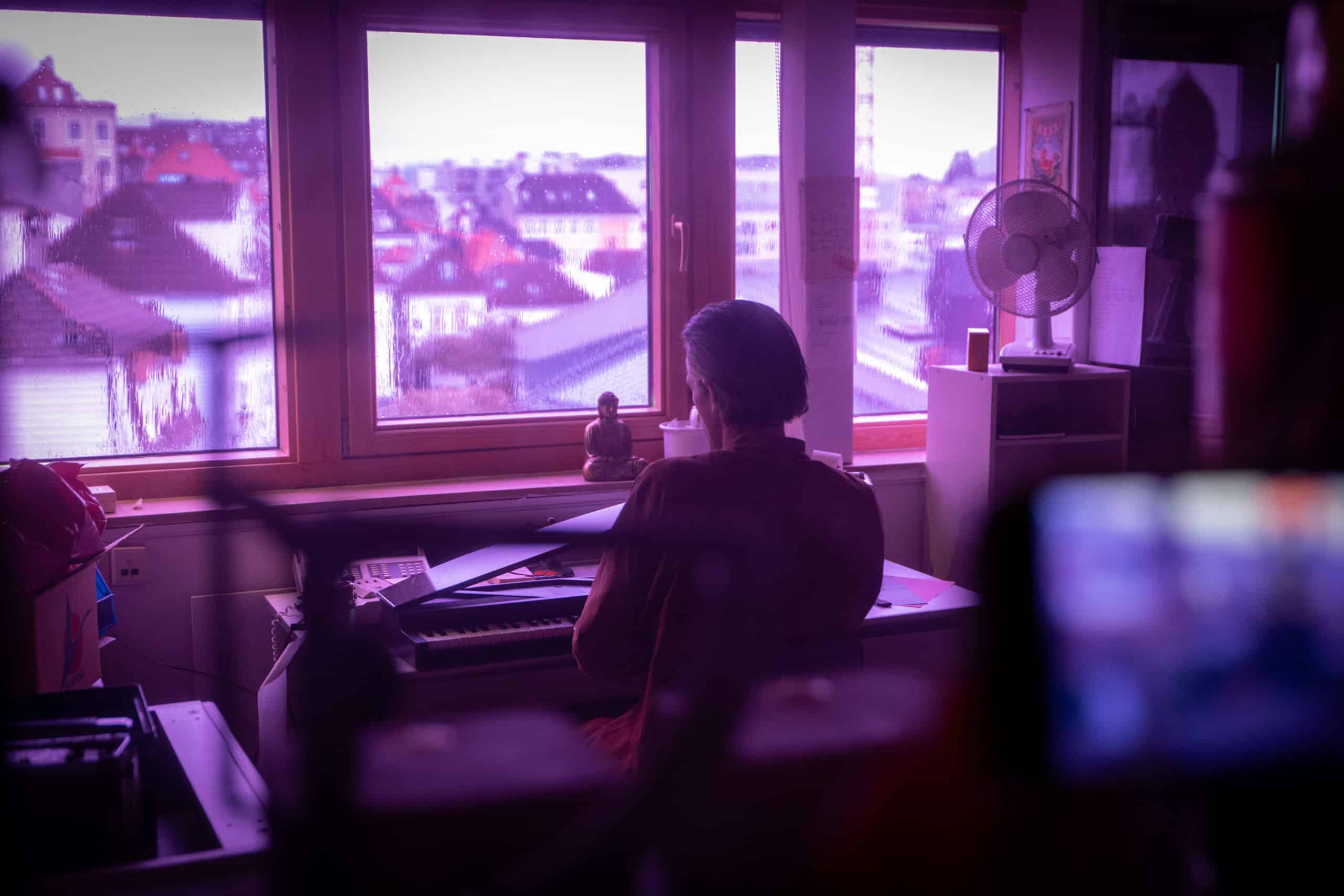 En person sitter ved et tastatur foran et vindu og ser ut på et bylandskap i rosa belysning.