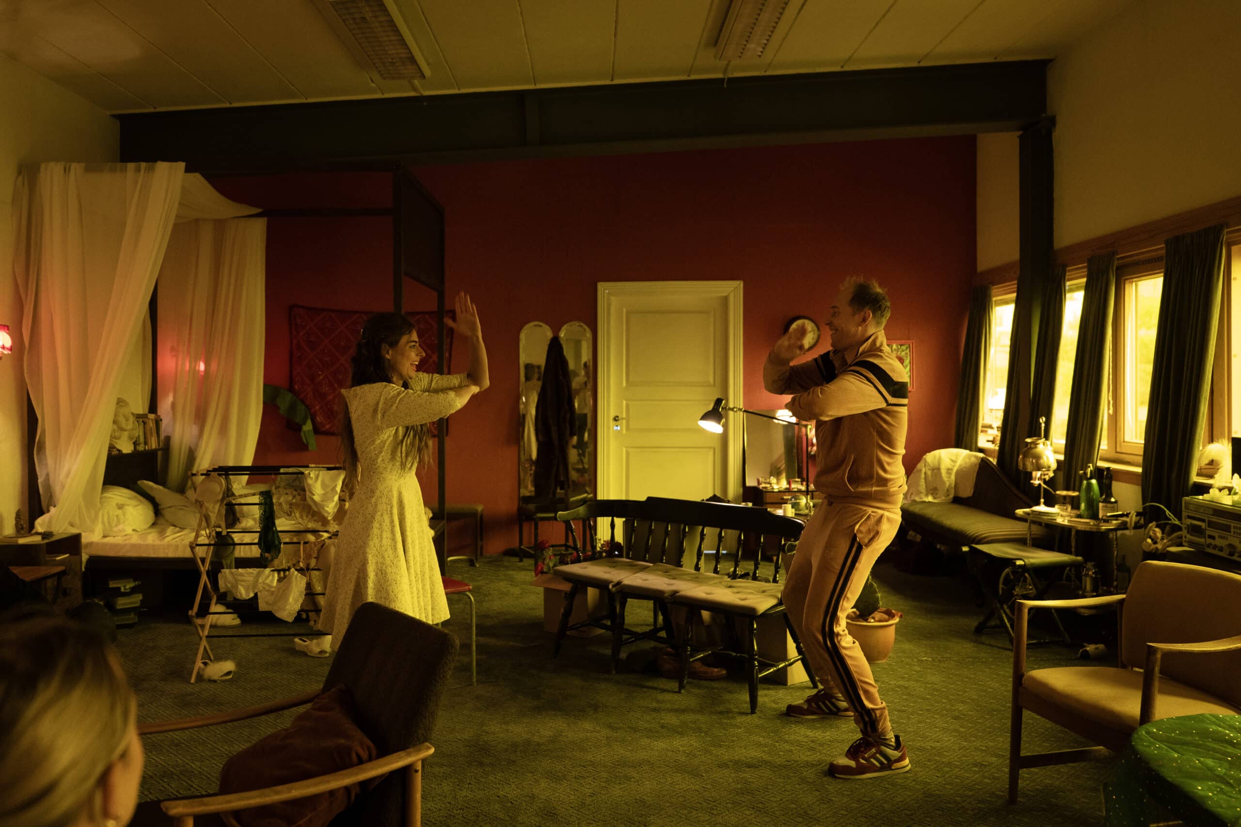 To personer står vendt mot hverandre i et svakt opplyst rom, den ene med hendene hevet, den andre holder en gjenstand. Rommet er fullt av senger, stoler og ulike husholdningsartikler.