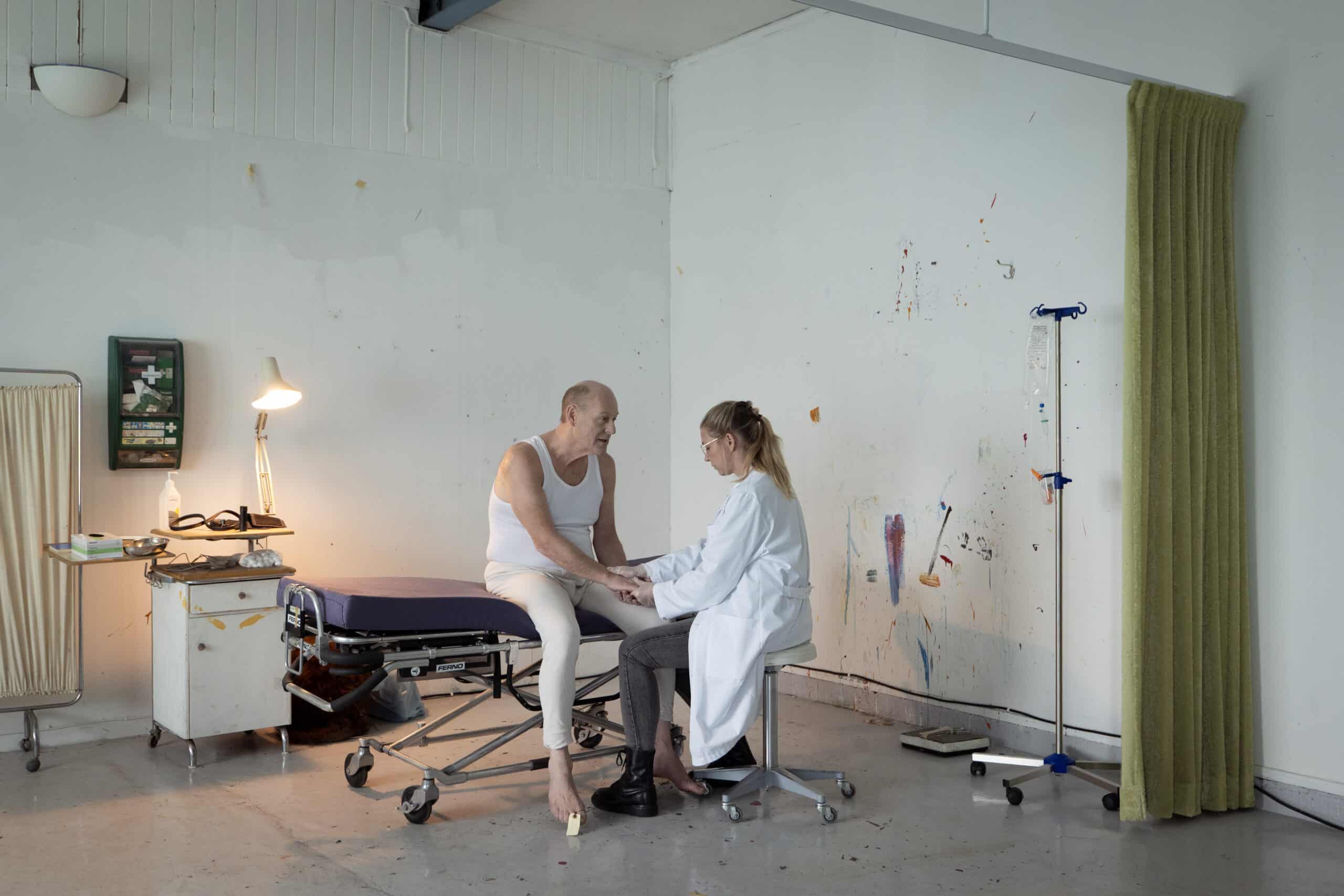 En eldre mann i tanktop sitter på en sykehusseng med bandasjerte ben, mens en lege i hvit frakk holder hendene hans i et nakent, uoversiktlig medisinrom.