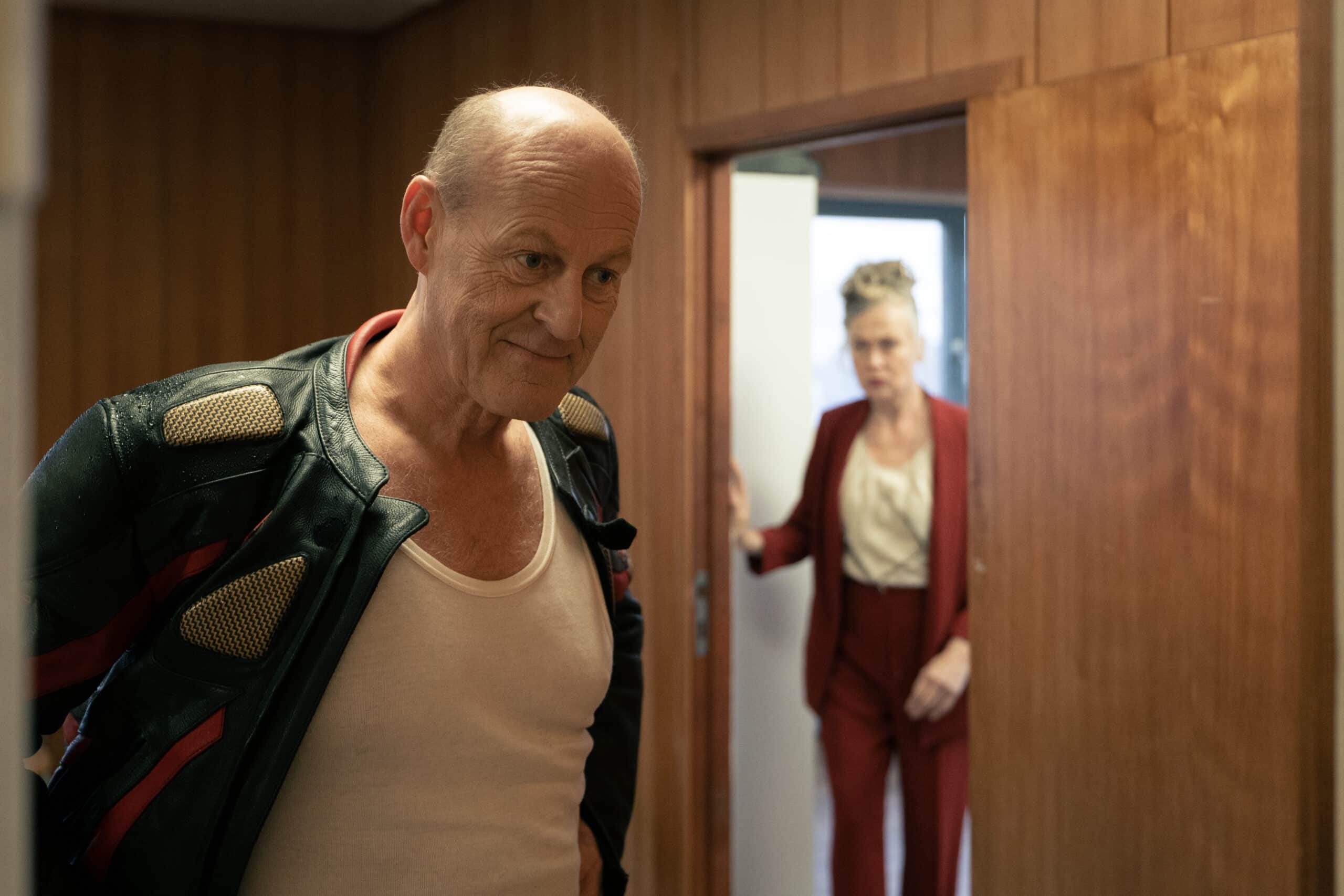 En skallet mann i skinnjakke og hvit tanktop står i forgrunnen, mens en kvinne i rød dress kommer inn i rommet bak ham gjennom en åpen dør.