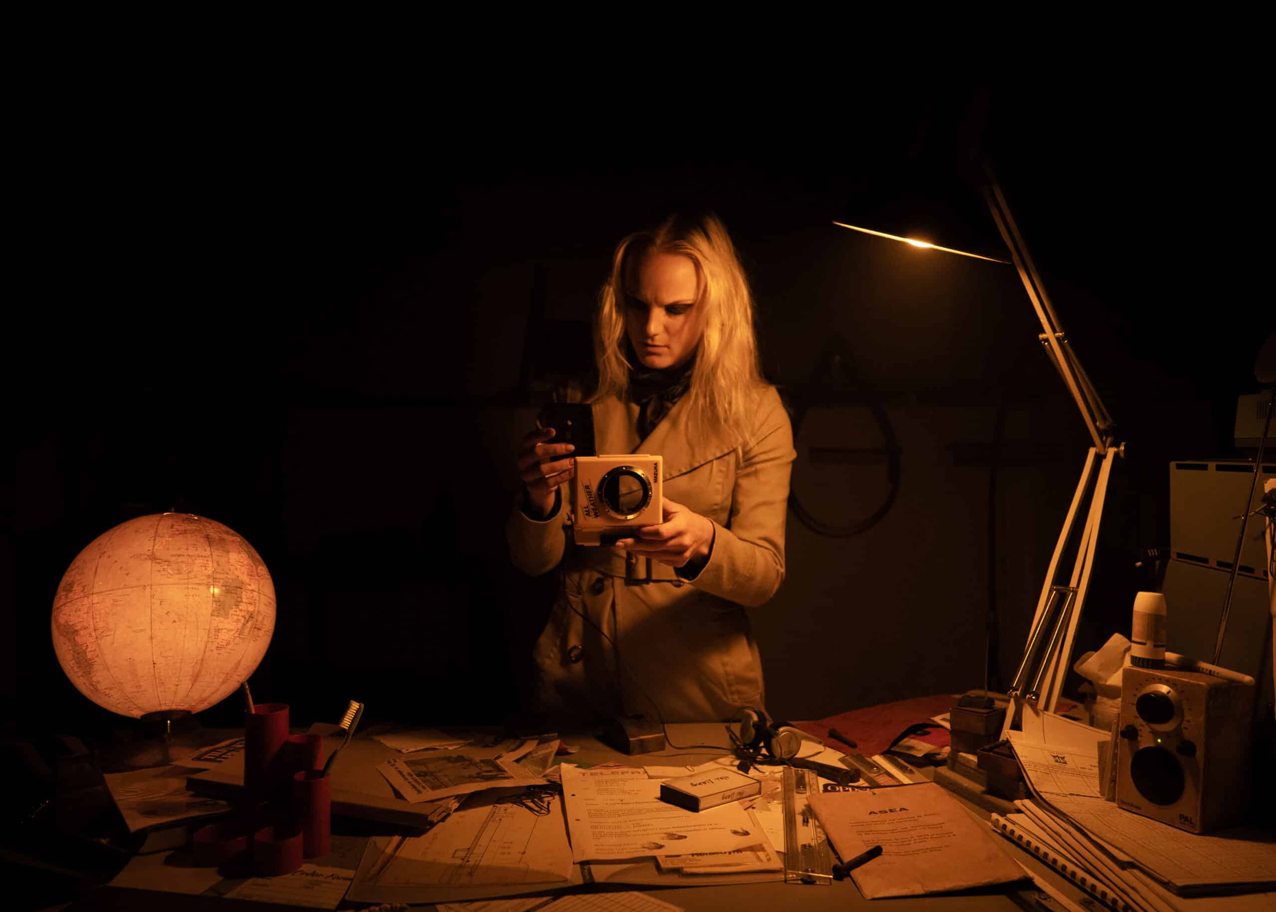En person med langt, blondt hår undersøker et vitenskapelig instrument ved et rotete skrivebord under en lampe, med dokumenter, en globus og utstyr spredt utover arbeidsområdet.