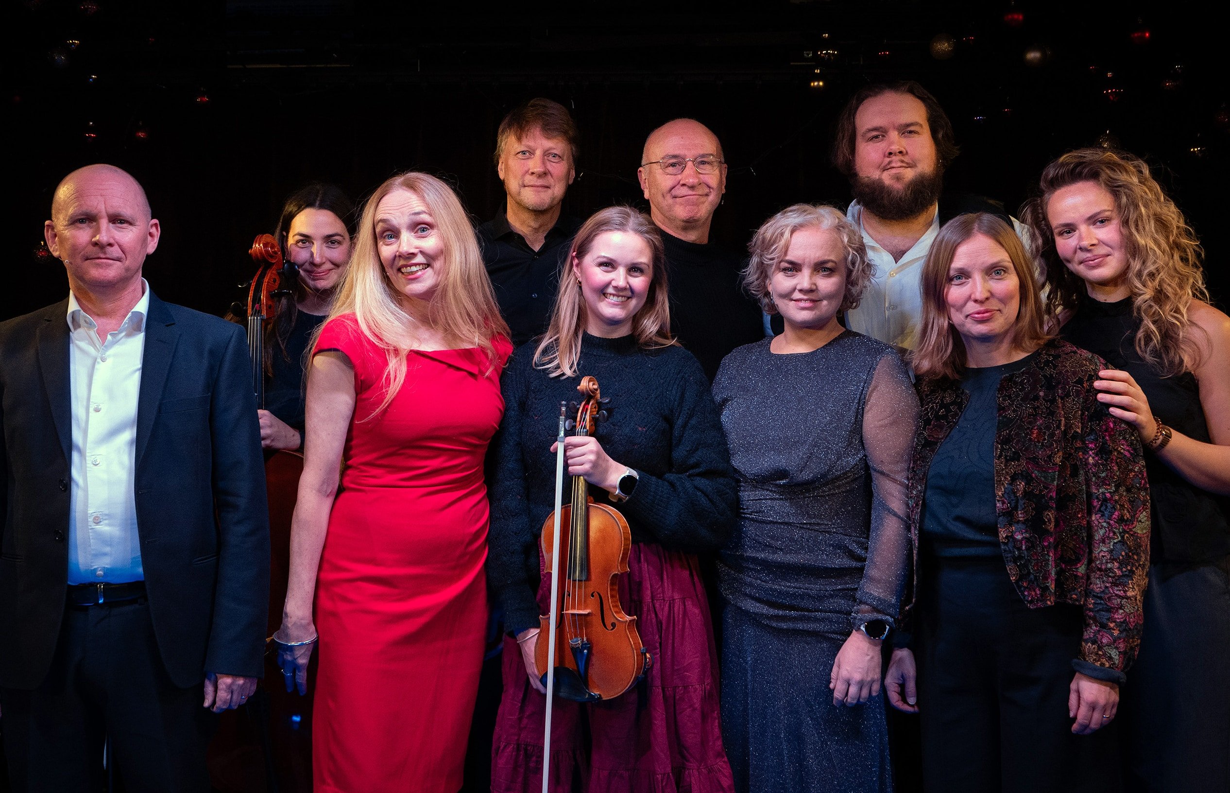 En gruppe på ti voksne, noen med musikkinstrumenter i hendene, poserer sammen for et bilde på scenen mot en mørk bakgrunn.