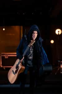 En person i hettejakke holder en akustisk gitar og en gitarkoffert på en svakt opplyst scene med spotlights og utstyr i bakgrunnen.