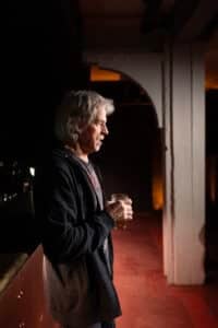 En eldre mann med grått hår står innendørs med et ølglass i hånden og ser til siden i et dunkelt opplyst rom.