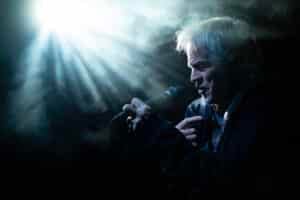 En eldre mann synger i en mikrofon under dramatisk scenelys med tåke og lysstråler i bakgrunnen.