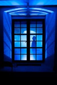 En silhuett av en person som står bak et vindu, belyst av blått lys, med skygger som kastes på veggene.