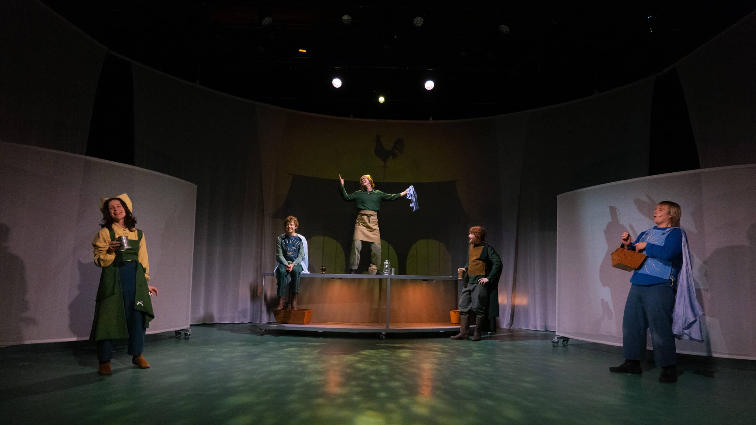 Fem skuespillere opptrer på en scene med buede, hvite fondplater; én står på et bord med armene hevet, mens de andre poserer rundt dem i teatralsk belysning.