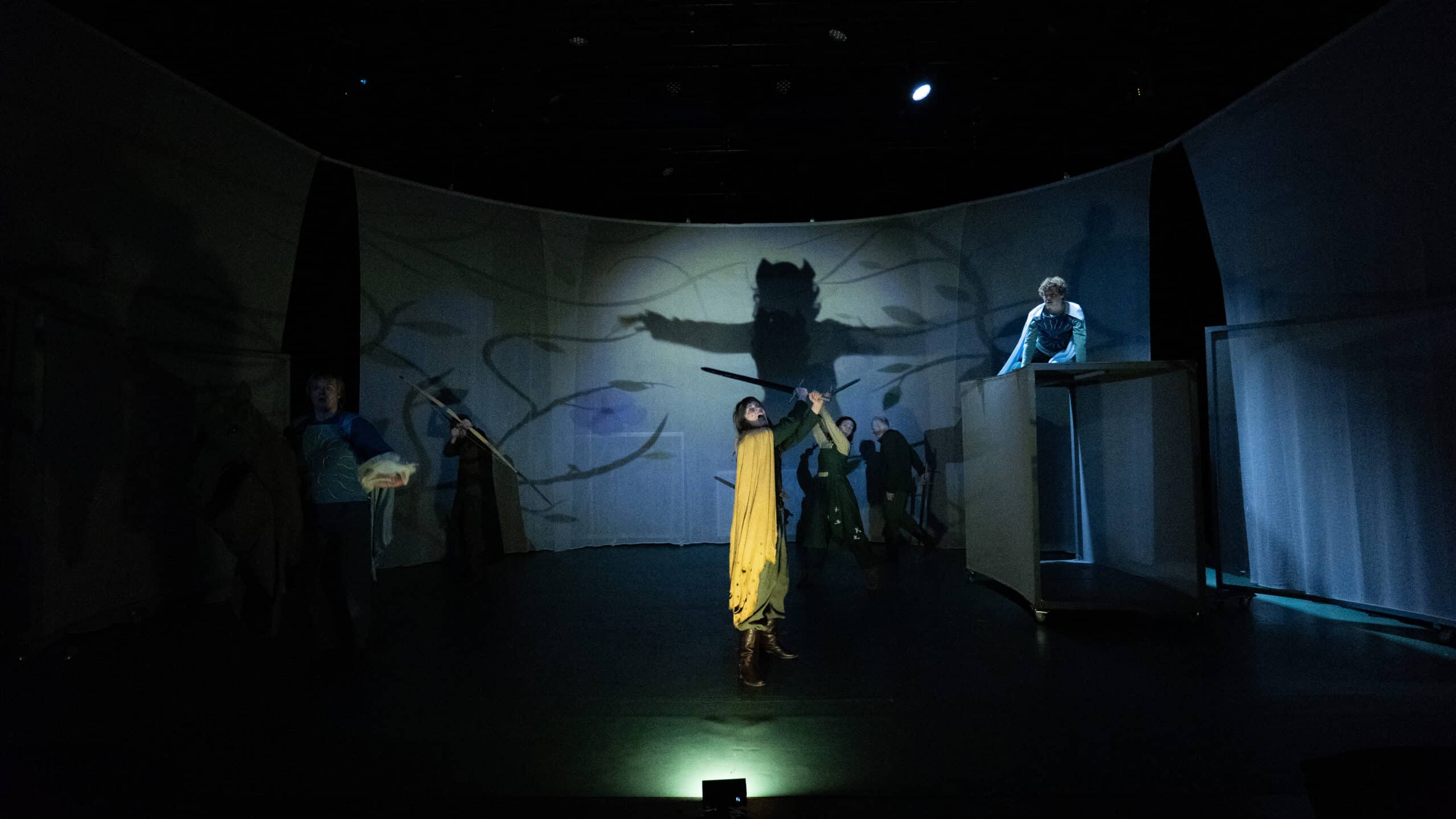 En scene med skuespillere i dramatiske positurer, en i gult kostyme midt på scenen, med en stor skyggeaktig figur projisert på et bakteppe og svak blå belysning.