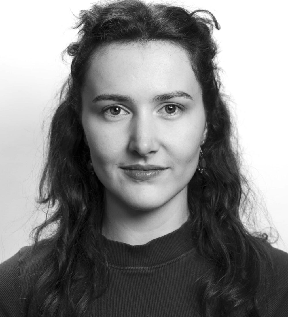 Svart-hvitt portrett av en kvinne med bølgende mørkt hår, iført langermet topp, som ser direkte inn i kameraet med et nøytralt uttrykk.