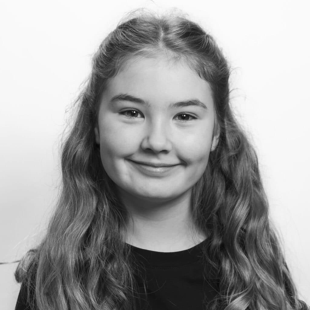 En ung jente med langt, bølget hår og et lite smil står vendt mot kameraet. Hun har på seg en mørk skjorte, og bakgrunnen er ensfarget og lys. Bildet er i svart-hvitt.