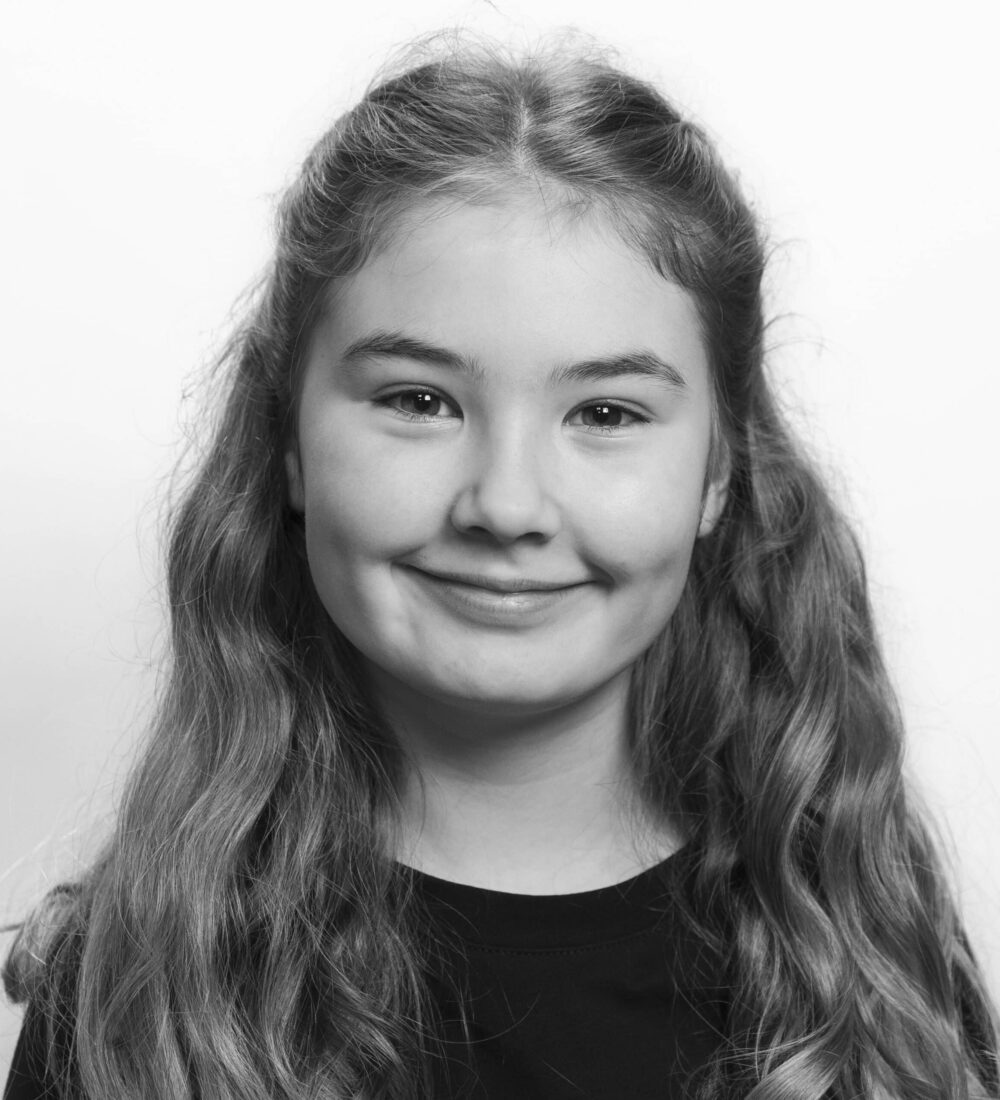 En ung jente med langt, bølget hår og et lite smil står vendt mot kameraet. Hun har på seg en mørk skjorte, og bakgrunnen er ensfarget og lys. Bildet er i svart-hvitt.