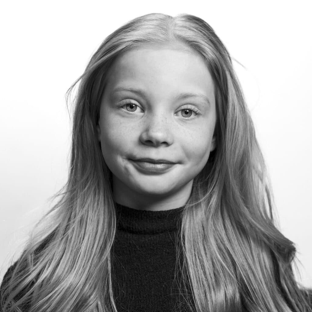 Svart-hvitt portrett av en ung jente med langt hår, iført en mørk topp, som ser inn i kameraet med et nøytralt uttrykk.