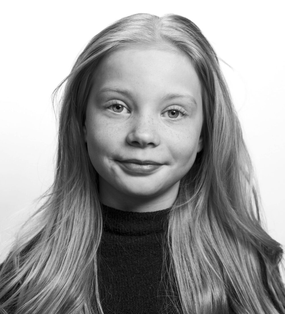 Svart-hvitt portrett av en ung jente med langt hår, iført en mørk topp, som ser inn i kameraet med et nøytralt uttrykk.