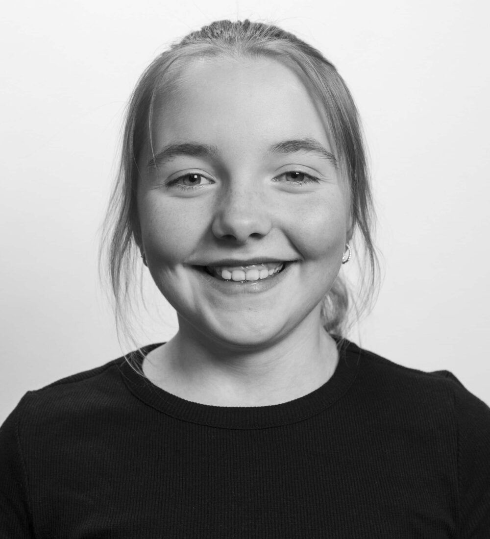 En ung person med langt hår smiler mot kameraet, iført en mørk topp mot en lys bakgrunn.