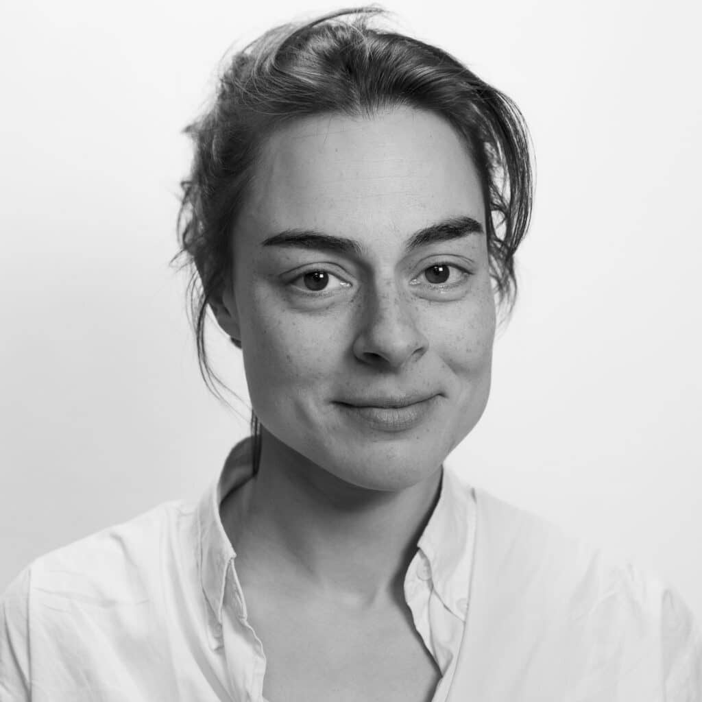 En kvinne med lys hud og mørkt hår som er bakoverbøyd, iført hvit skjorte, ser på kameraet med et lite smil mot en vanlig hvit bakgrunn.
