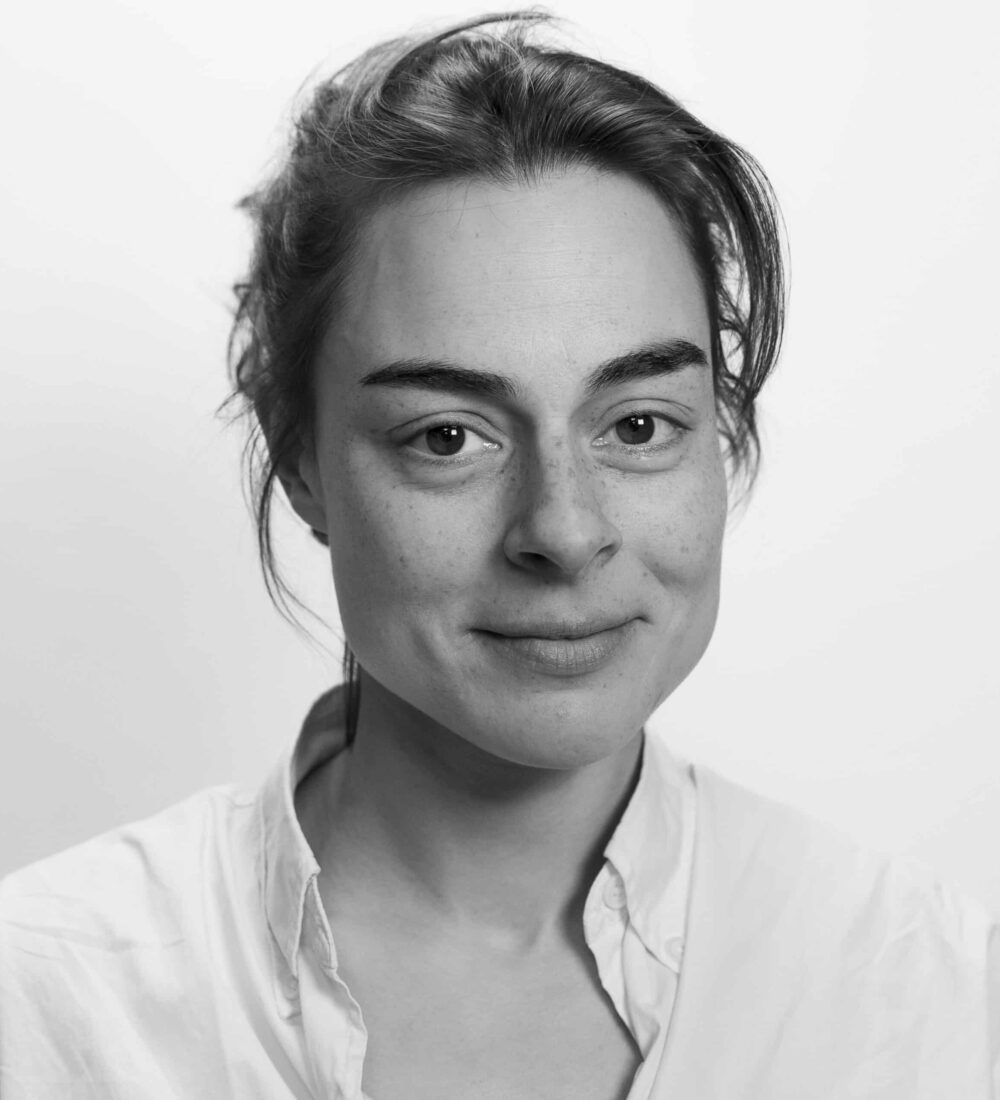 En kvinne med lys hud og mørkt hår som er bakoverbøyd, iført hvit skjorte, ser på kameraet med et lite smil mot en vanlig hvit bakgrunn.