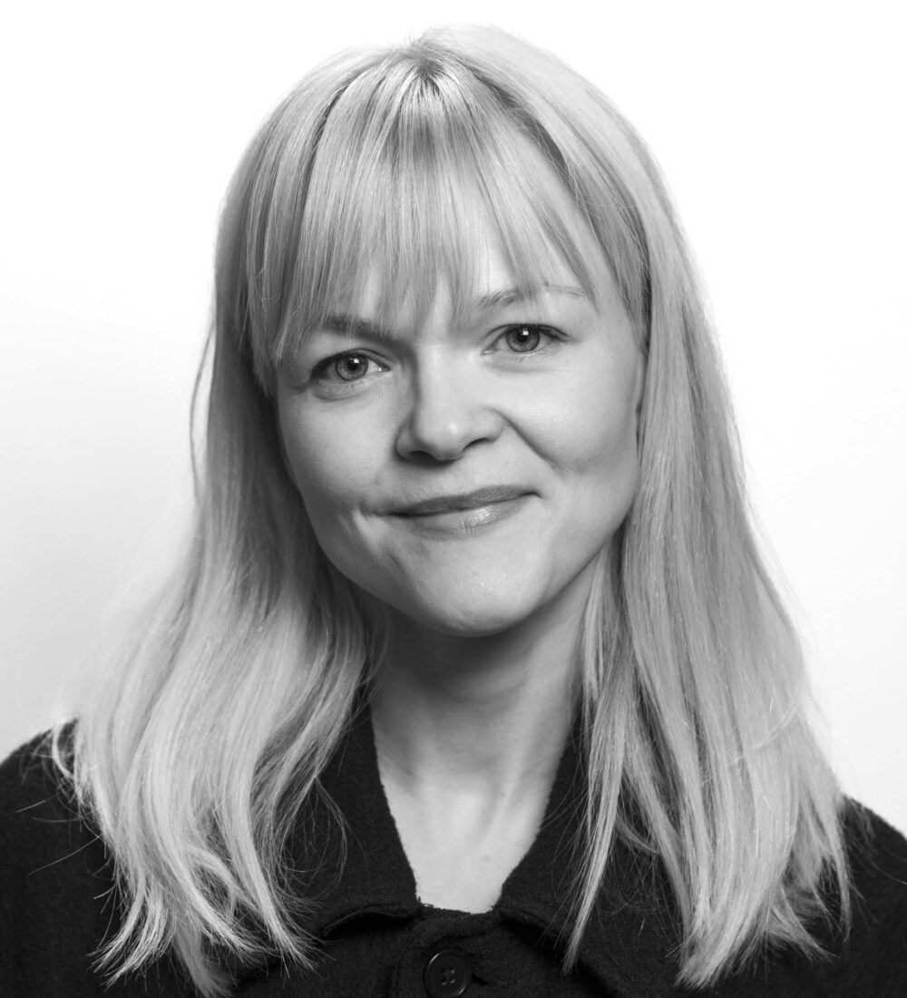 En kvinne med rett, lyst hår og lugg smiler lett, iført mørk jakke, i et svart-hvitt portrett med ensfarget bakgrunn.