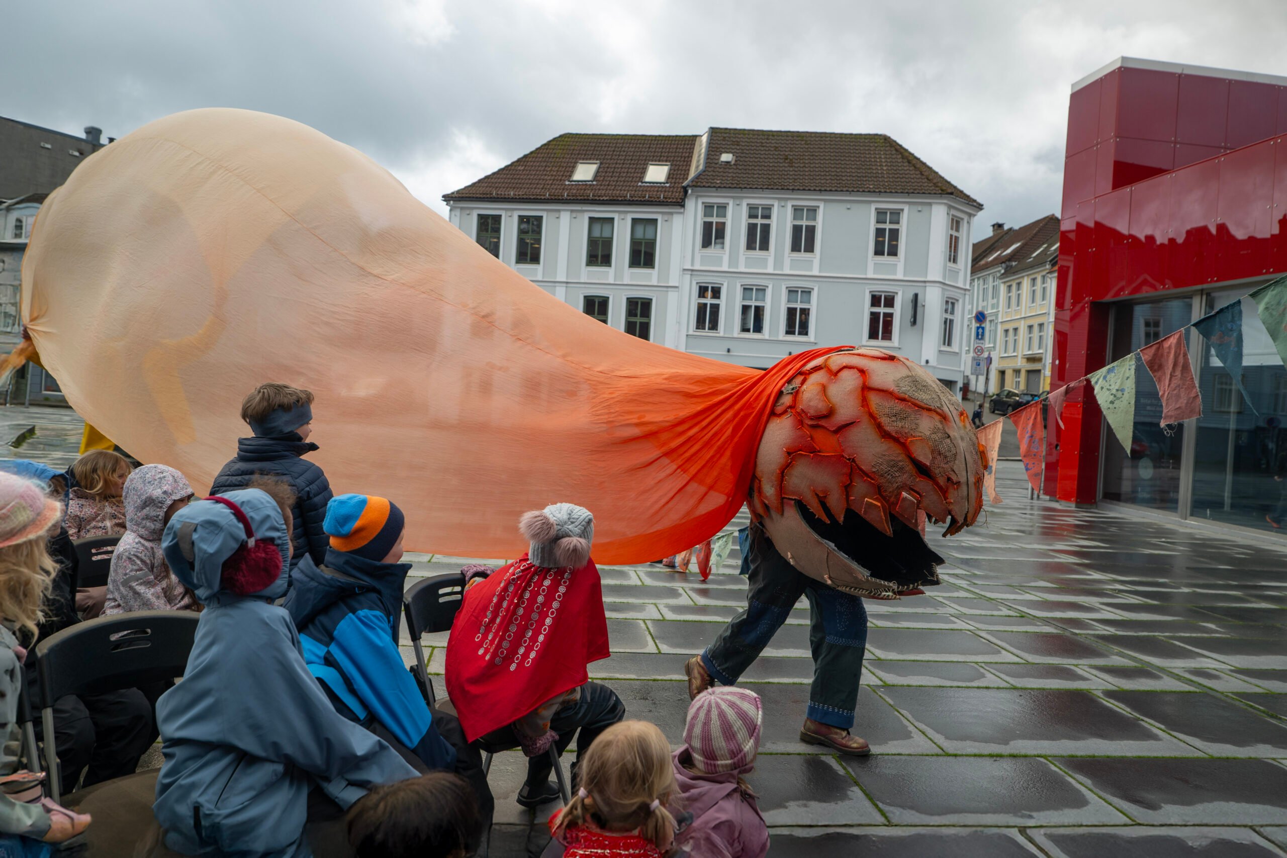 En artist i et stort, oransje og beige skapningskostyme går forbi sittende barn utendørs på et torg en overskyet dag.