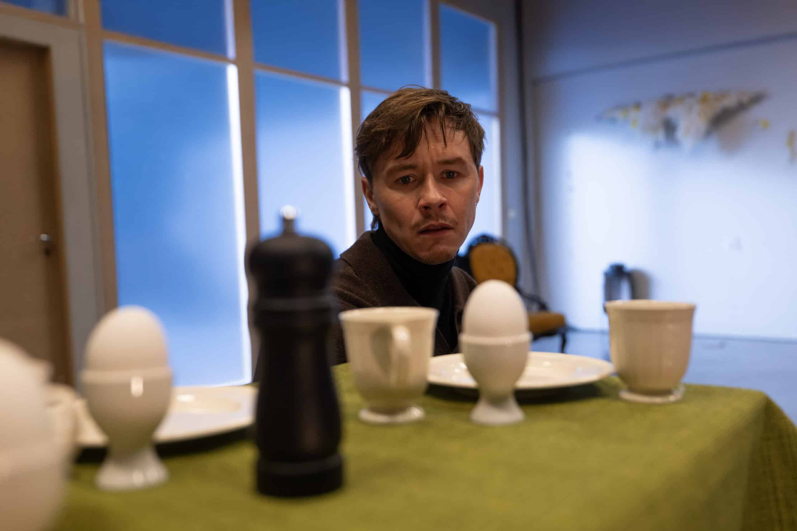 En mann sitter ved et bord dekket med egg, kopper, tallerkener og en pepperkvern og ser fremover med et nøytralt ansiktsuttrykk.