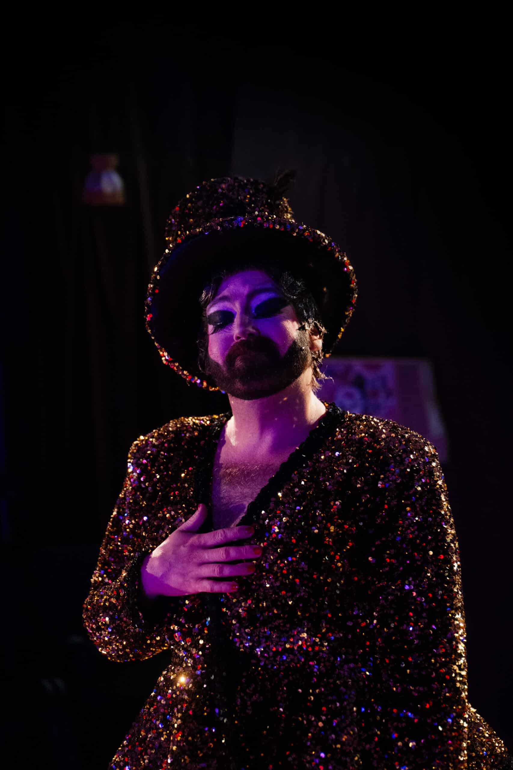 En person i dramatisk scenemakeup og glitrende kostyme med matchende hatt står med den ene hånden på brystet under teatralsk belysning.