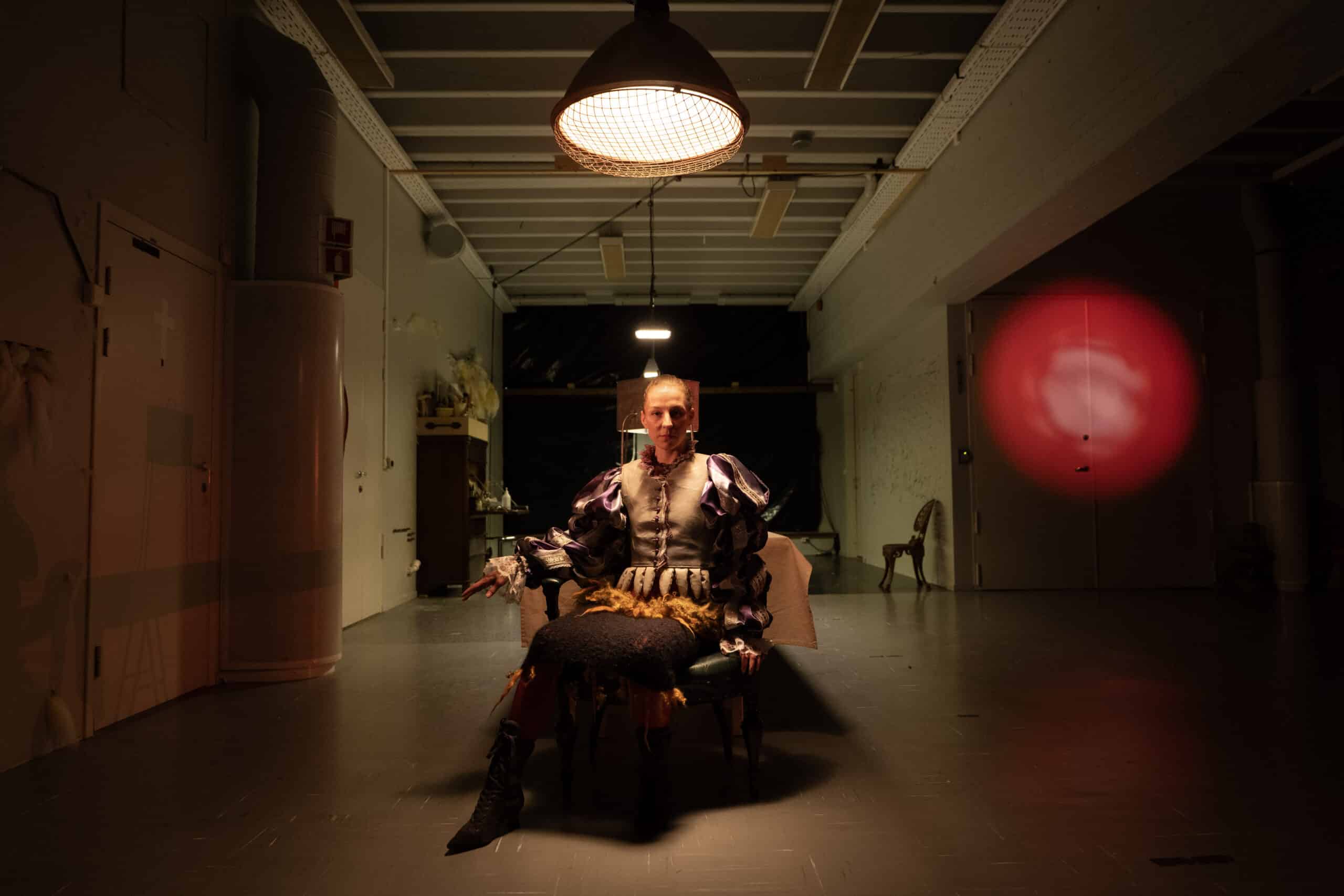 En person i teaterkostyme sitter på en stol under en spotlight i et svakt opplyst rom med industrielle trekk og rødt lys på veggen.