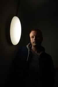 En mann i et mørkt rom står ved siden av en lys, rund vegglampe, som kaster skarpe skygger på ansiktet hans og veggen bak ham.