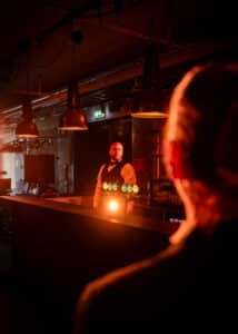 En bartender står bak en bar opplyst av oransje lys og ser mot en person i forgrunnen, hvis ansiktstrekk er uskarpe og ute av fokus. Man kan se industriell innredning og pendellamper.