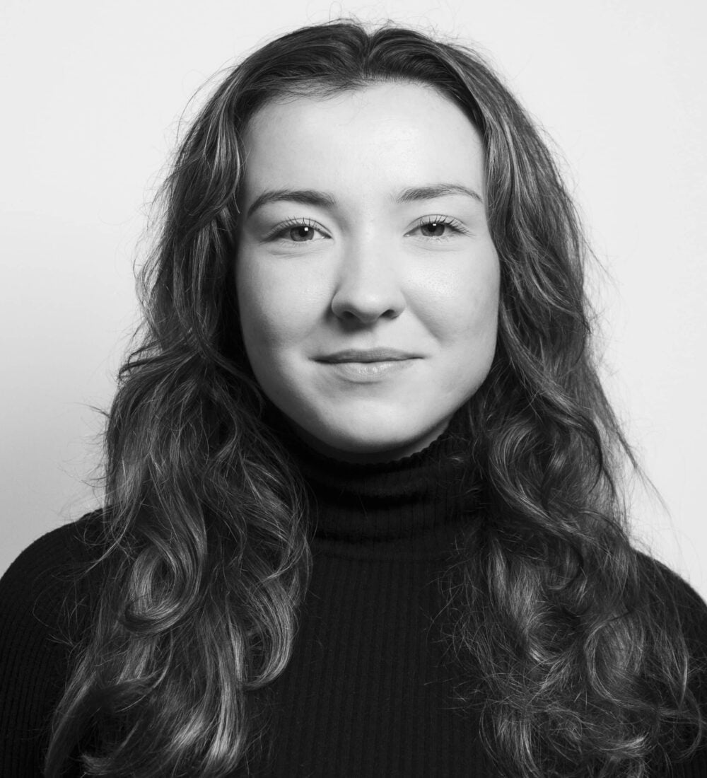 En kvinne med langt, bølget hår og en mørk genser med høyhals vender seg mot kameraet og smiler svakt mot en ensfarget, lys bakgrunn.
