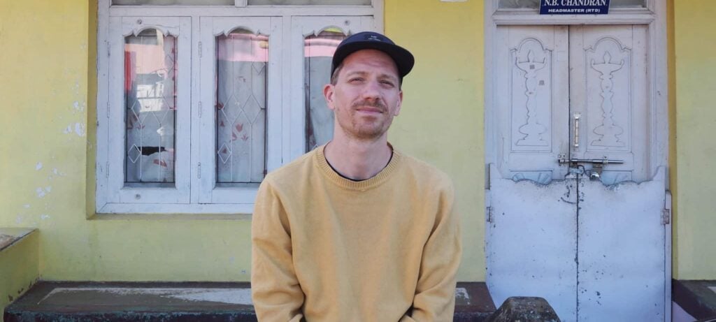 En mann i gul genser og svart lue lener seg mot en lav vegg foran en gul bygning med hvite vinduer og en dør merket «K.R. Chandran».