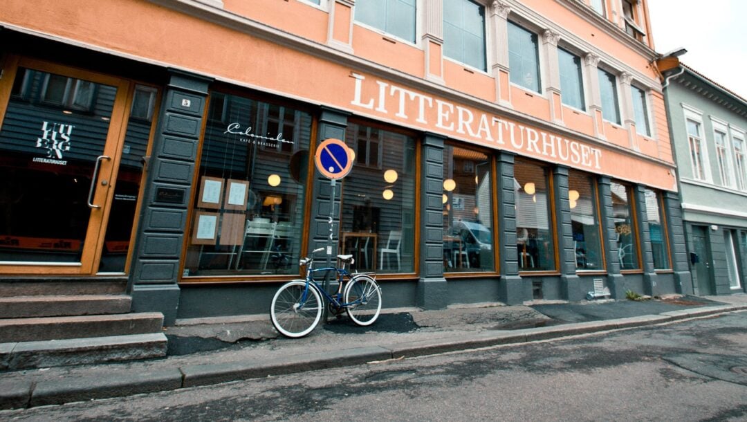 En sykkel står parkert utenfor en grå og oransje bygning med påskriften «LITTERATURHUSET» på fasaden. Det står et parkeringsforbudsskilt foran bygningen, og man kan se runde lys inne i bygningen.