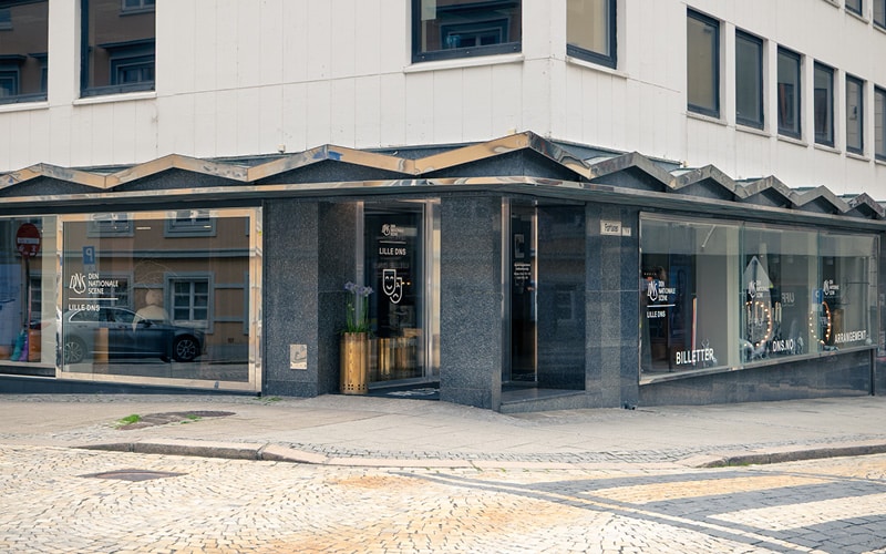 Gatebilde av en moderne hjørnebygning med store glassvinduer, mørk steinfasade og skilt for billetter og suvenirer på innsiden.