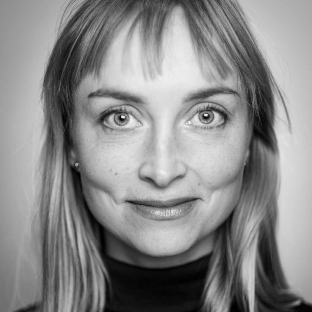 Svart-hvitt nærbilde av en kvinne med rett, lyst hår, lugg, lyse øyne og et lite smil, iført mørk topp, som ser direkte inn i kameraet.