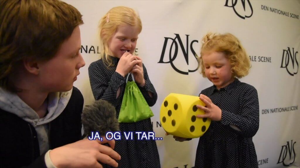 En person intervjuer to små barn som holder en stor skummatris, mot et bakteppe med logoer.